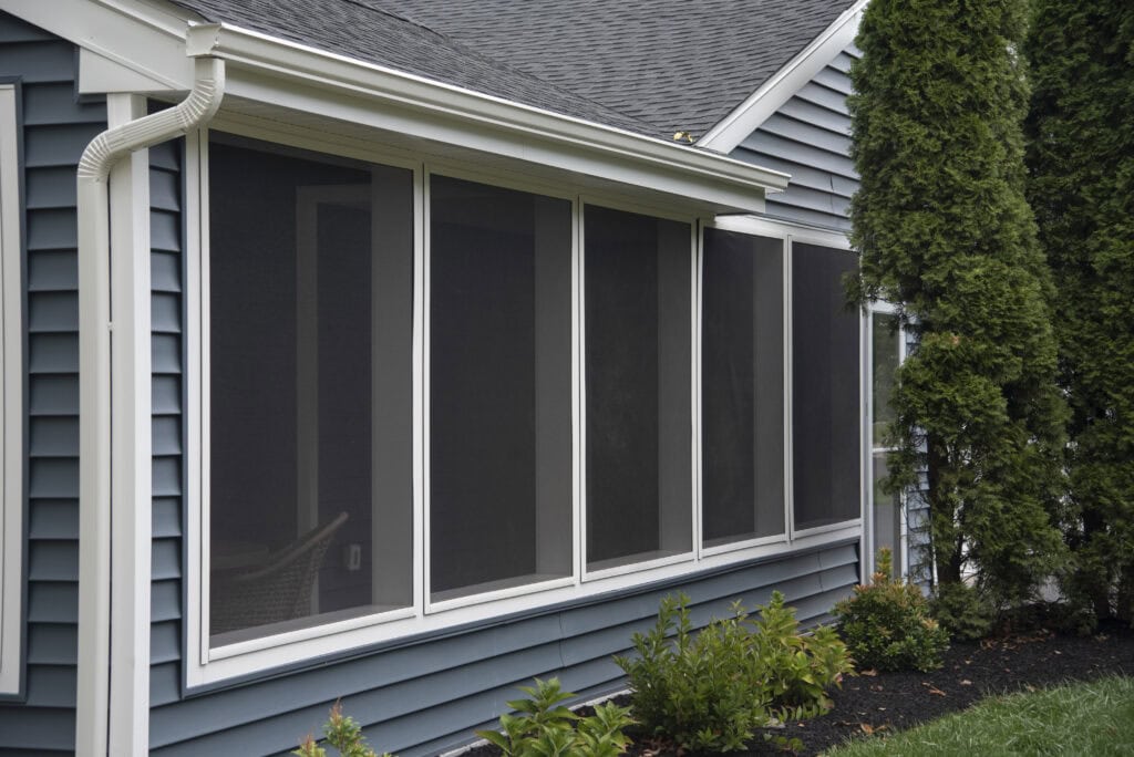A close-up view of a house with blue siding and large, screened-in porch windows, next to green shrubs and tall evergreen trees.