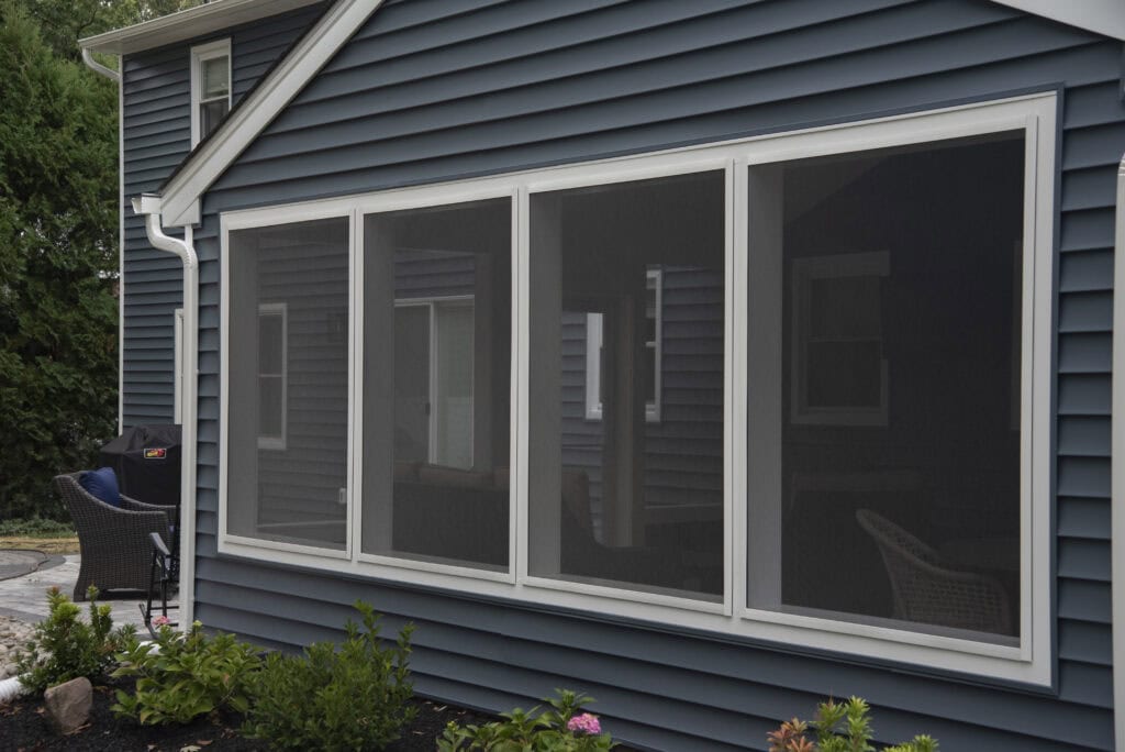 A screened-in porch attached to a blue house with white trim, featuring large screen panels and patio furniture visible through the screens. Small plants line the garden bed below the windows.