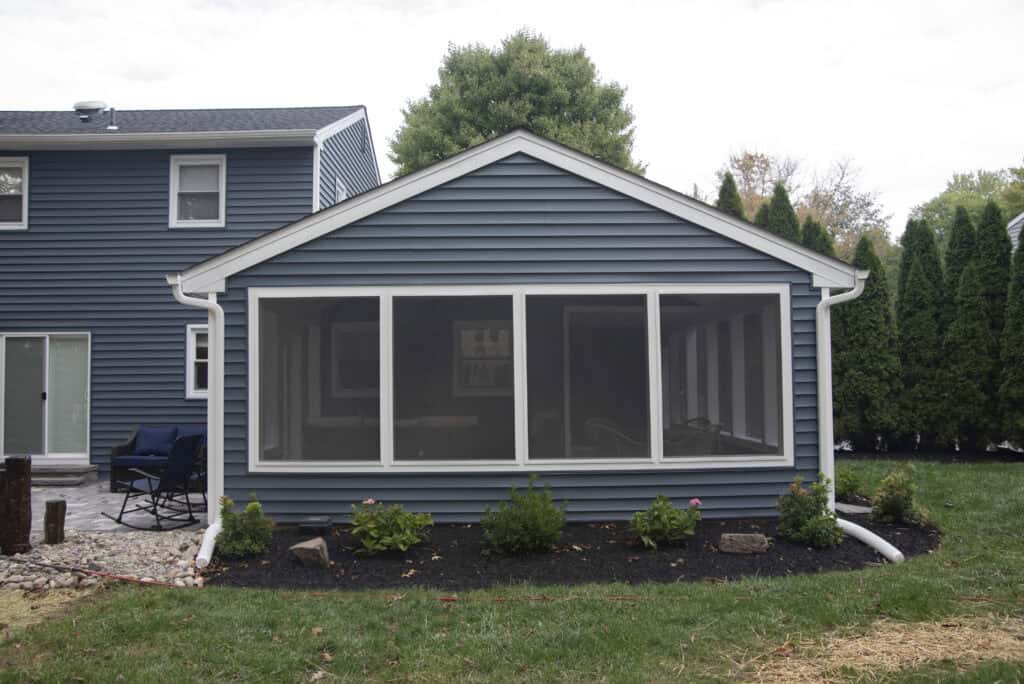 A blue house with a screened-in porch, white trim, and landscaped bushes in front; patio furniture sits to the left, and the lawn and trees surround the area.