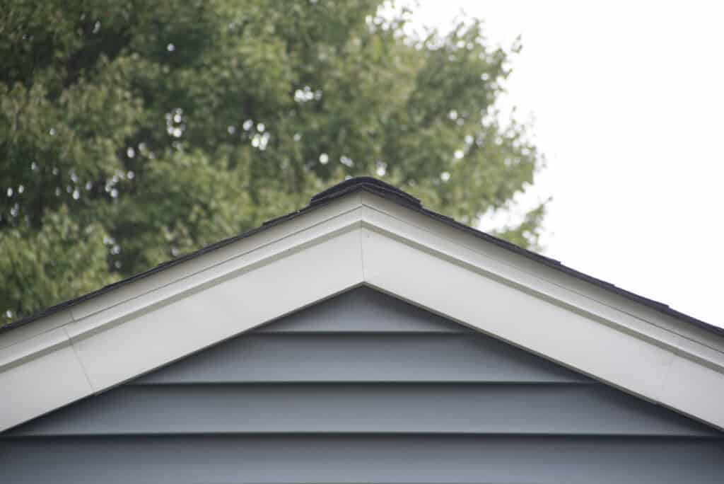 The peak of a house roof with gray siding and white trim, centered in the image, with green leafy trees blurred in the background.