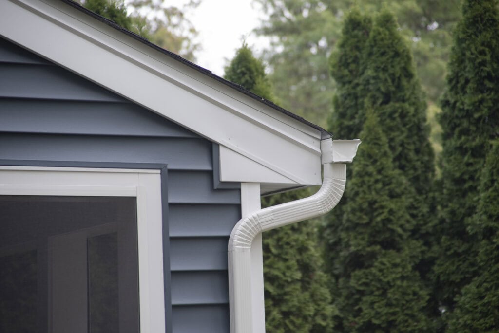 Close-up of a house corner with blue siding, a white rain gutter and downspout, and a screened porch area; tall green trees are visible in the background.