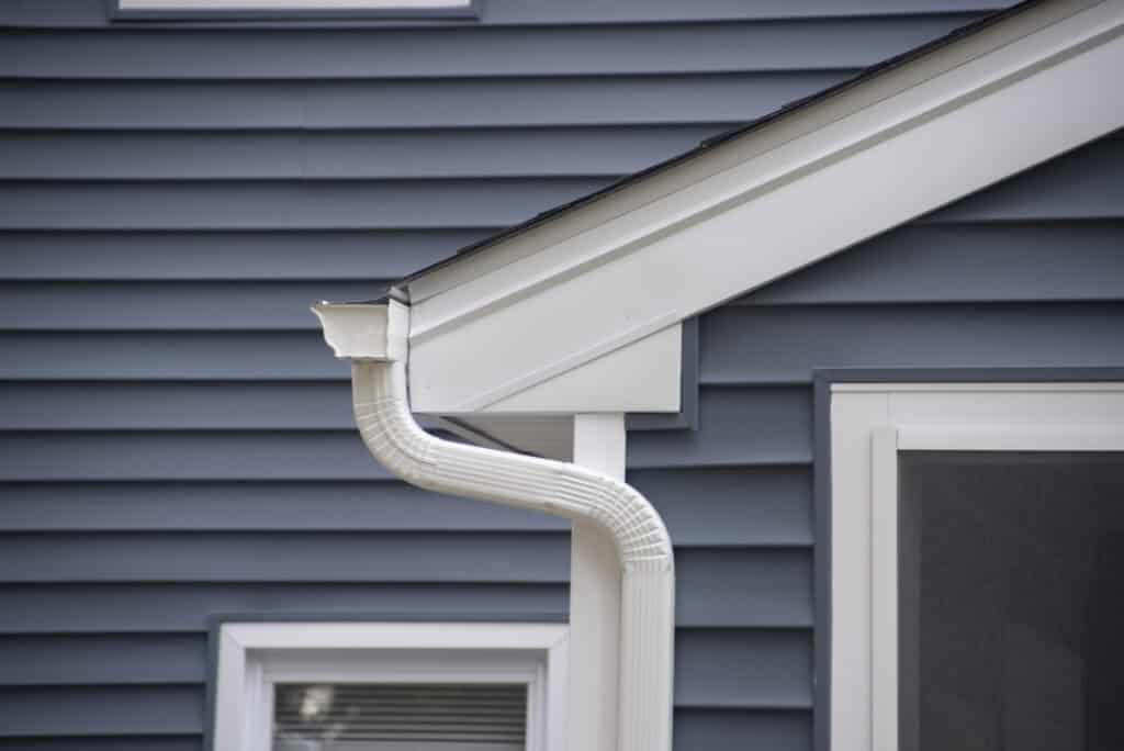 Close-up of a house corner with blue vinyl siding, white trim, and a white gutter with a downspout attached near the roof, above a window and a door.