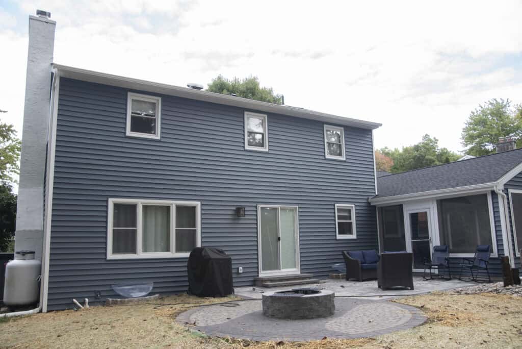 A two-story blue house with white trim, a patio area with outdoor furniture, a grill, and a circular fire pit in the backyard. Some grass is missing, revealing patches of bare ground.