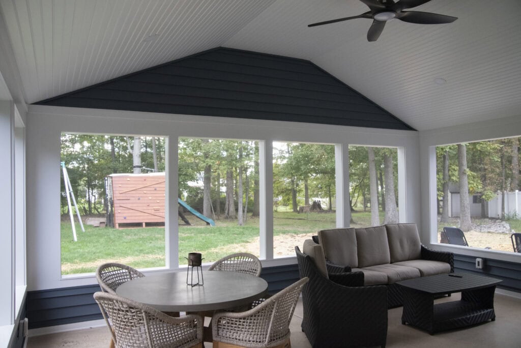 A bright sunroom with large windows, a round table with four wicker chairs, a cushioned sofa, and a coffee table. Outside, there is a grassy yard with trees and a wooden play structure.