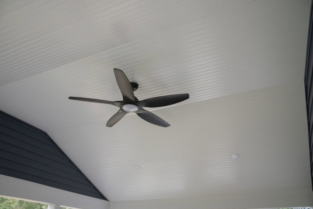 A modern black ceiling fan with five blades mounted on a white, beadboard-paneled, vaulted ceiling with two recessed lights and dark blue accent walls.