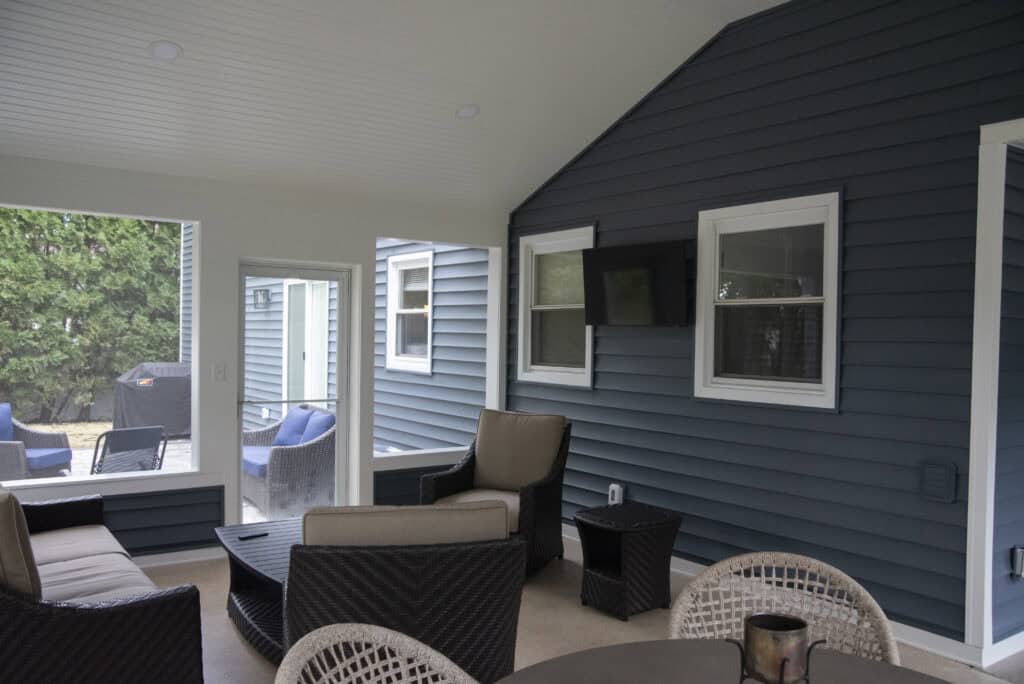 A screened-in porch with wicker patio furniture, a mounted TV, and blue siding walls. Large windows show an outdoor seating area and greenery. A round table and chairs are in the foreground.