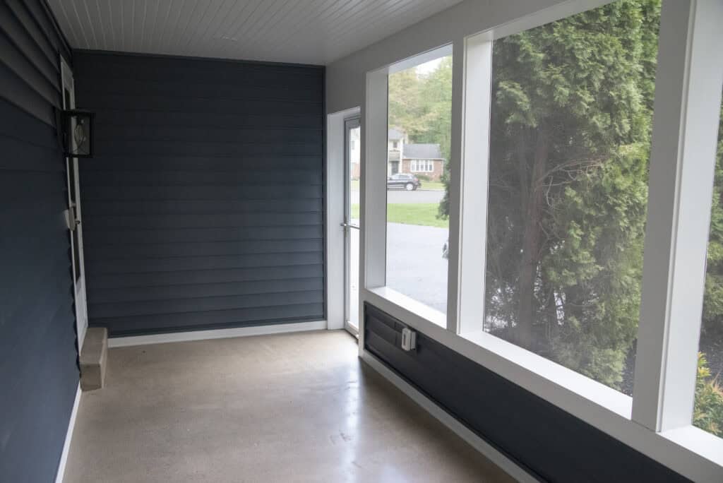 A covered porch with blue siding, large screened windows on one side, a small wall bench, and a door leading outside; a suburban street and greenery are visible through the screens.