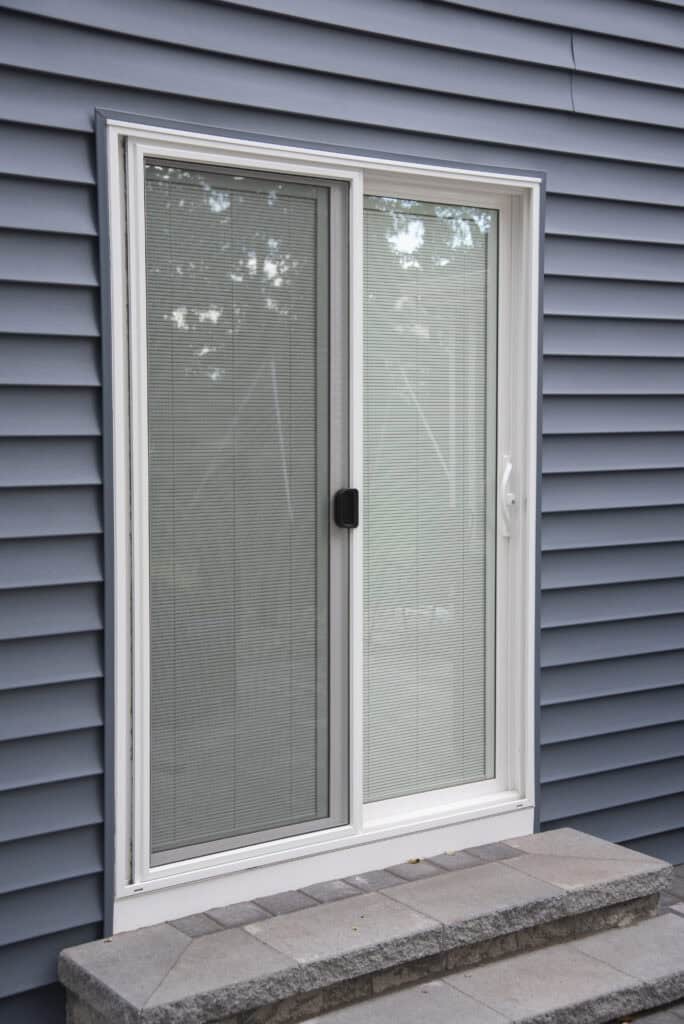 A white sliding glass door with built-in blinds, set in a blue-grey vinyl-sided exterior wall, with stone steps and a small landing in front of the door.