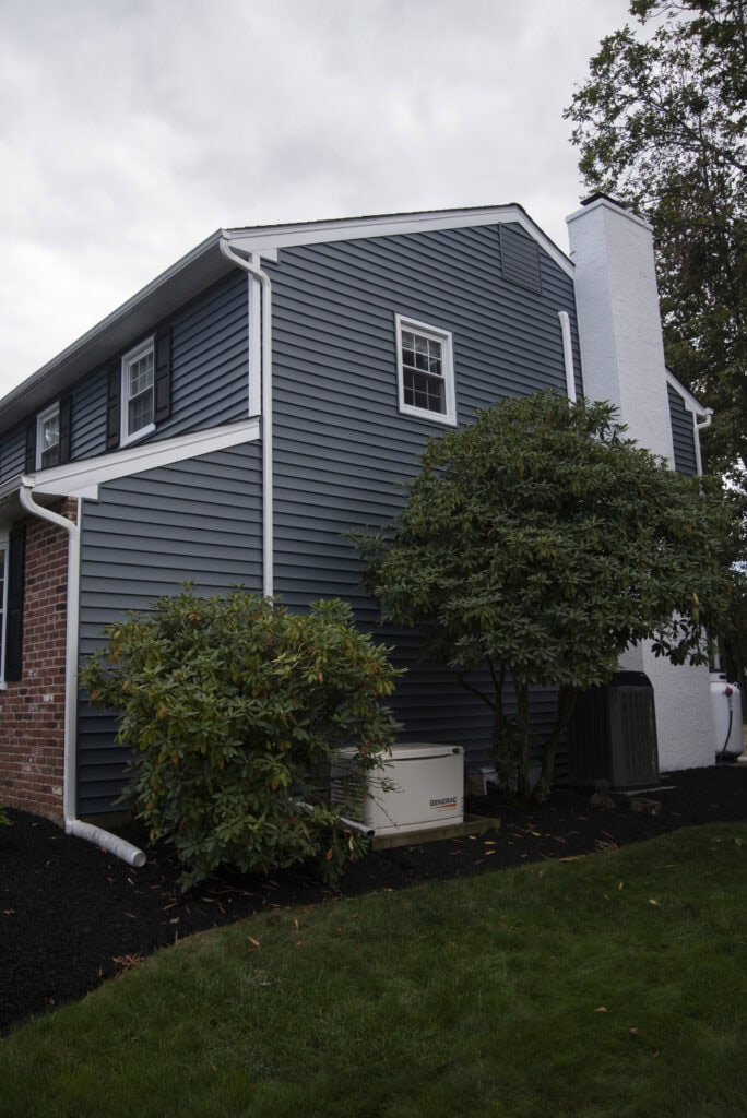 A two-story house with dark blue siding and white trim, a tall white chimney, green bushes, a generator, and an air conditioning unit beside it; overcast sky and green lawn in the foreground.