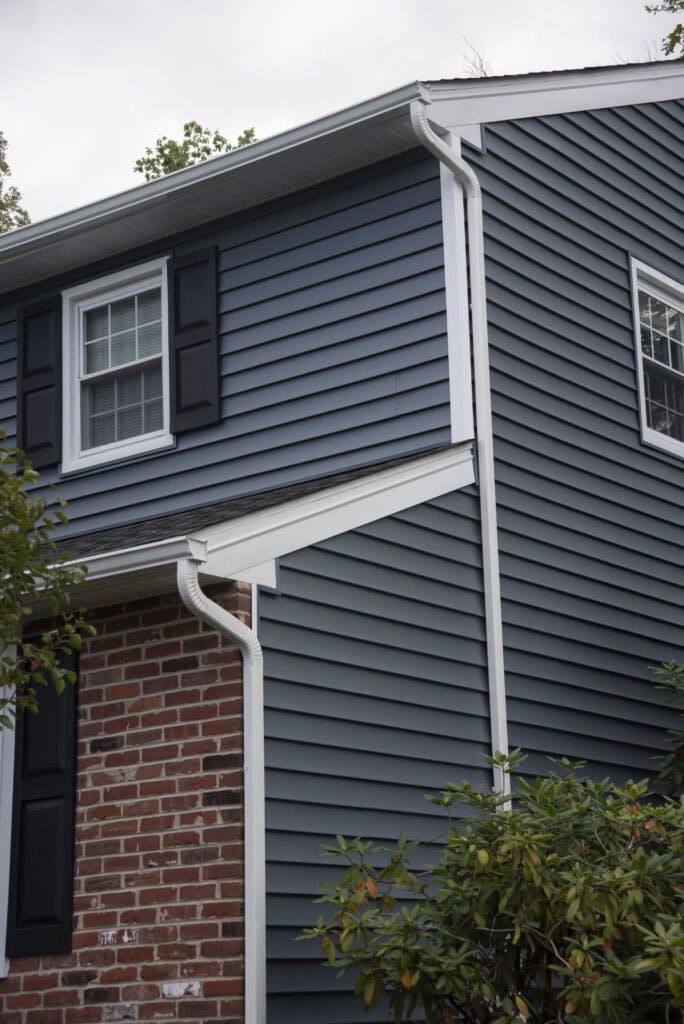 A close-up of a house exterior with blue vinyl siding, white trim, black shutters, and a red brick lower section beside green bushes.