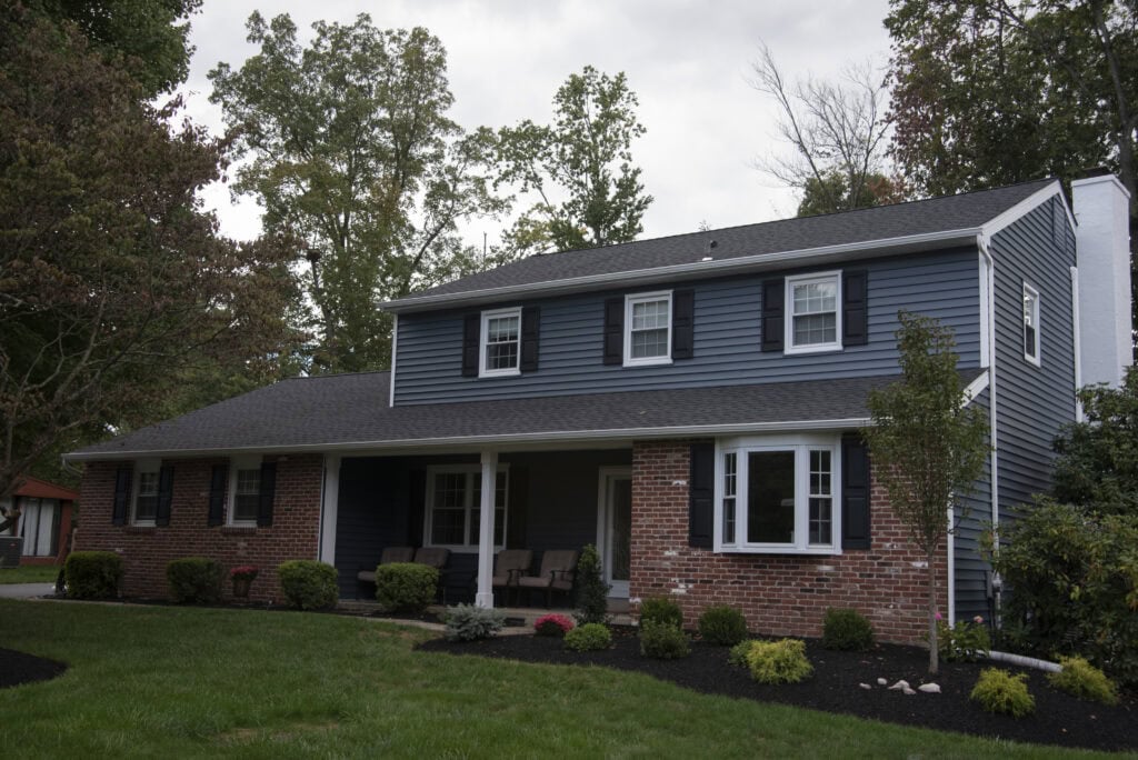 A two-story suburban house with blue siding on the upper floor and red brick on the lower, featuring white trim, a covered front porch with chairs, and a well-maintained lawn and shrubs in front.