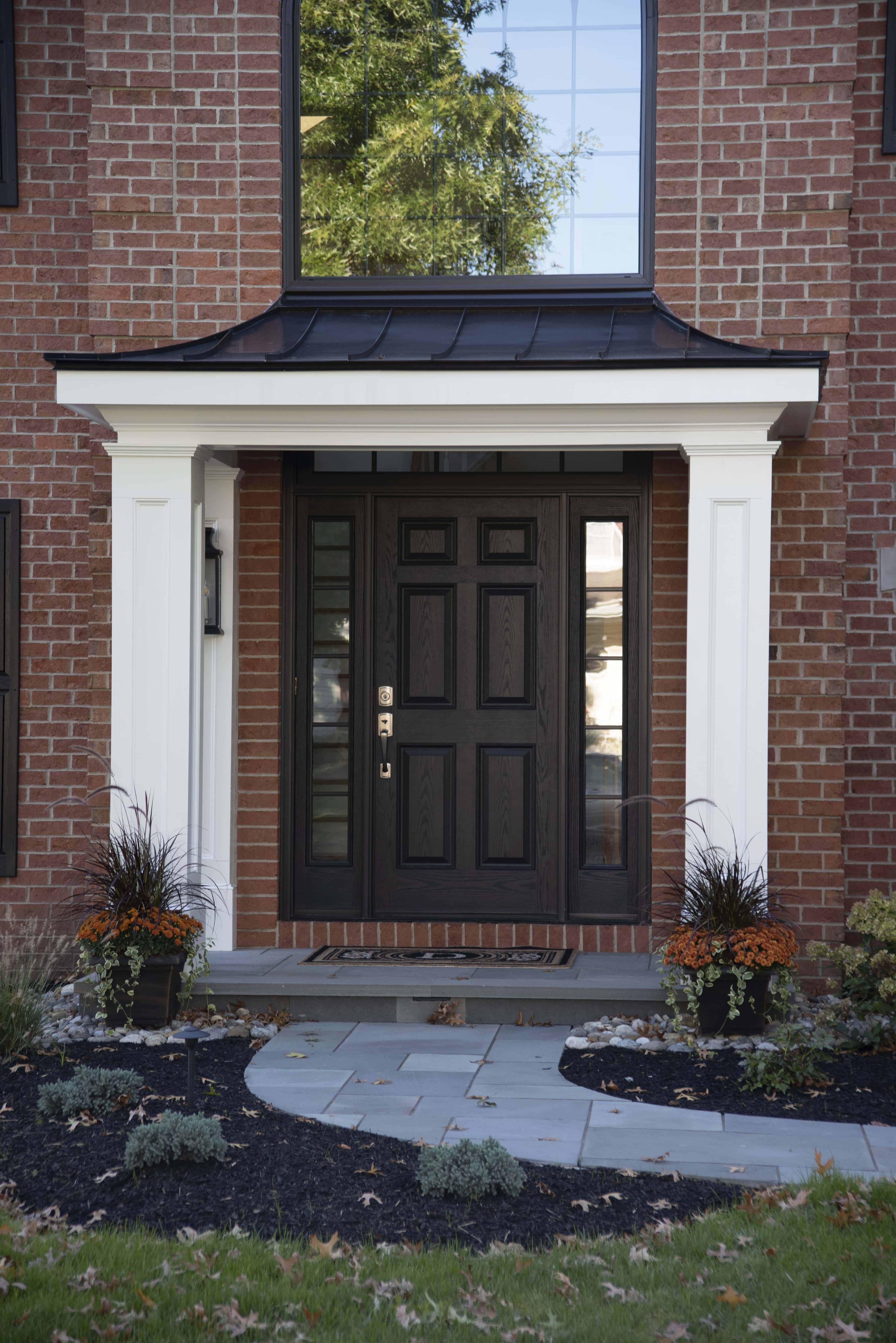 A modern brick house entrance with a black door, white trim, and sidelights. Two potted plants with fall flowers sit on either side of the door, and a curved stone path leads to the entry.