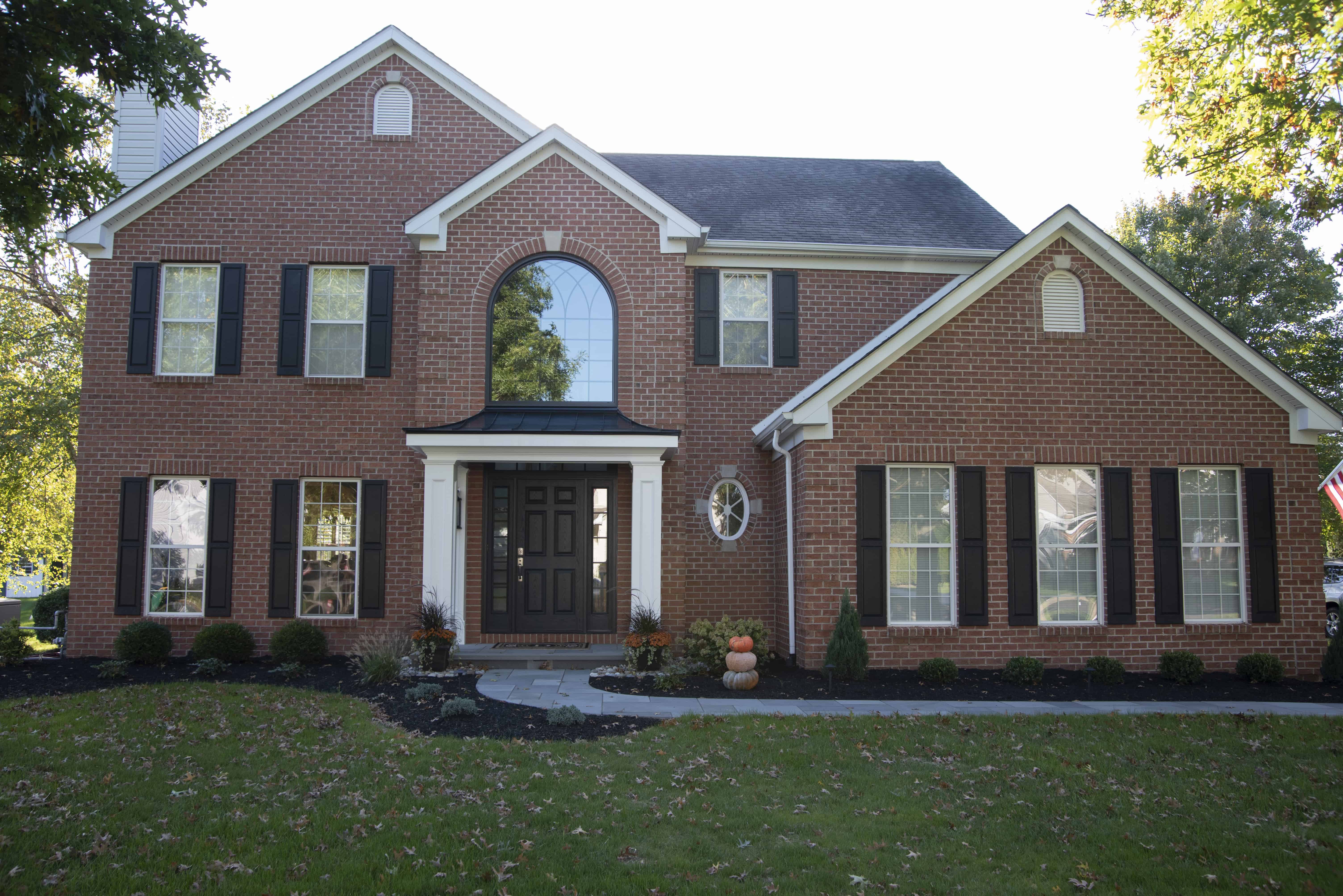 Two-story red brick house with black shutters, dark double front doors, arched window above the entrance, and a neatly landscaped front yard with green grass and autumn decorations near the entrance.