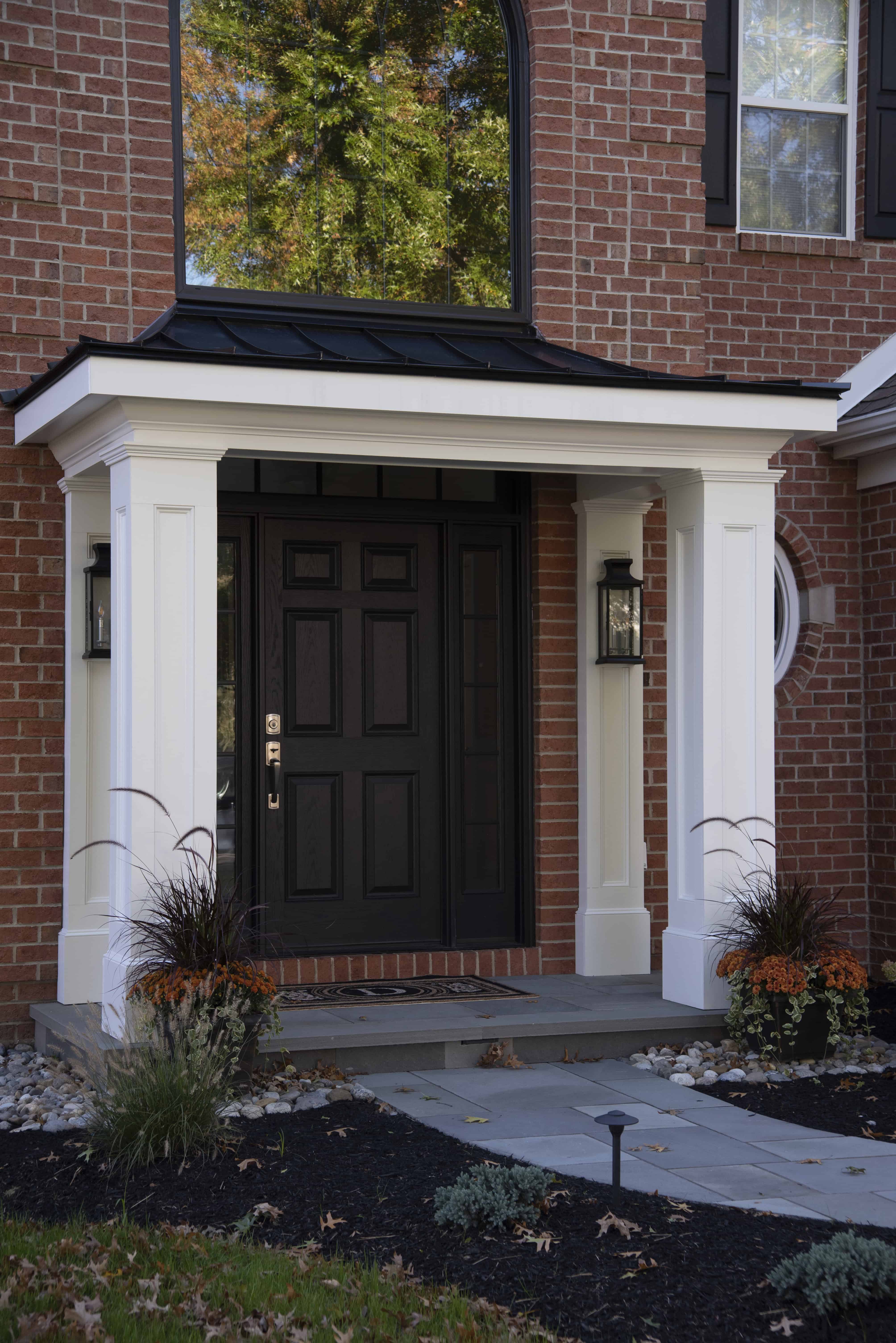 A brick house entryway with a black door, white columns, and a small porch. Two black lantern lights flank the door, and planters with grasses and orange flowers sit on either side of the steps.