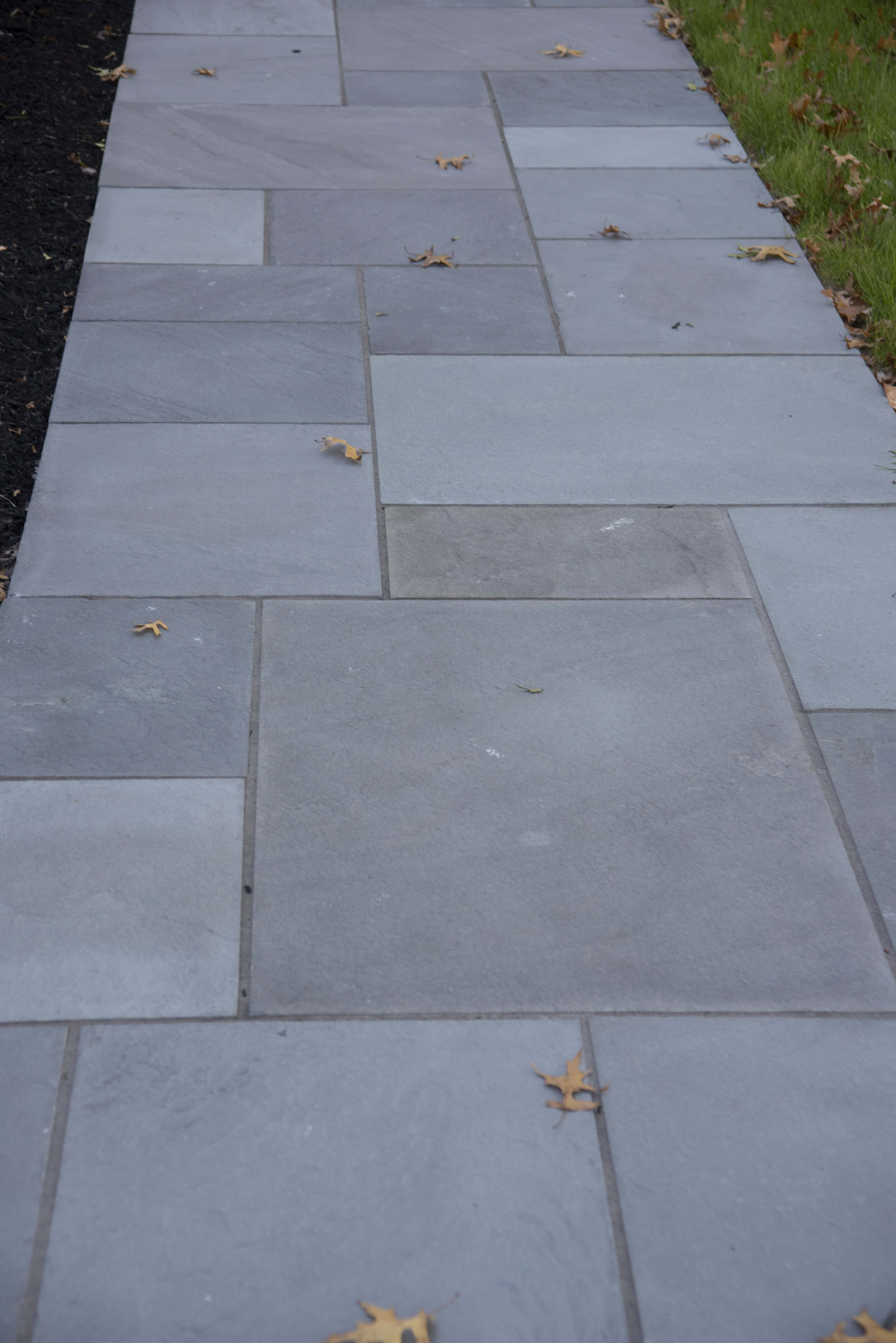 A stone walkway made of large gray rectangular and square tiles, bordered by black mulch on one side and green grass on the other, with scattered brown leaves on the path.