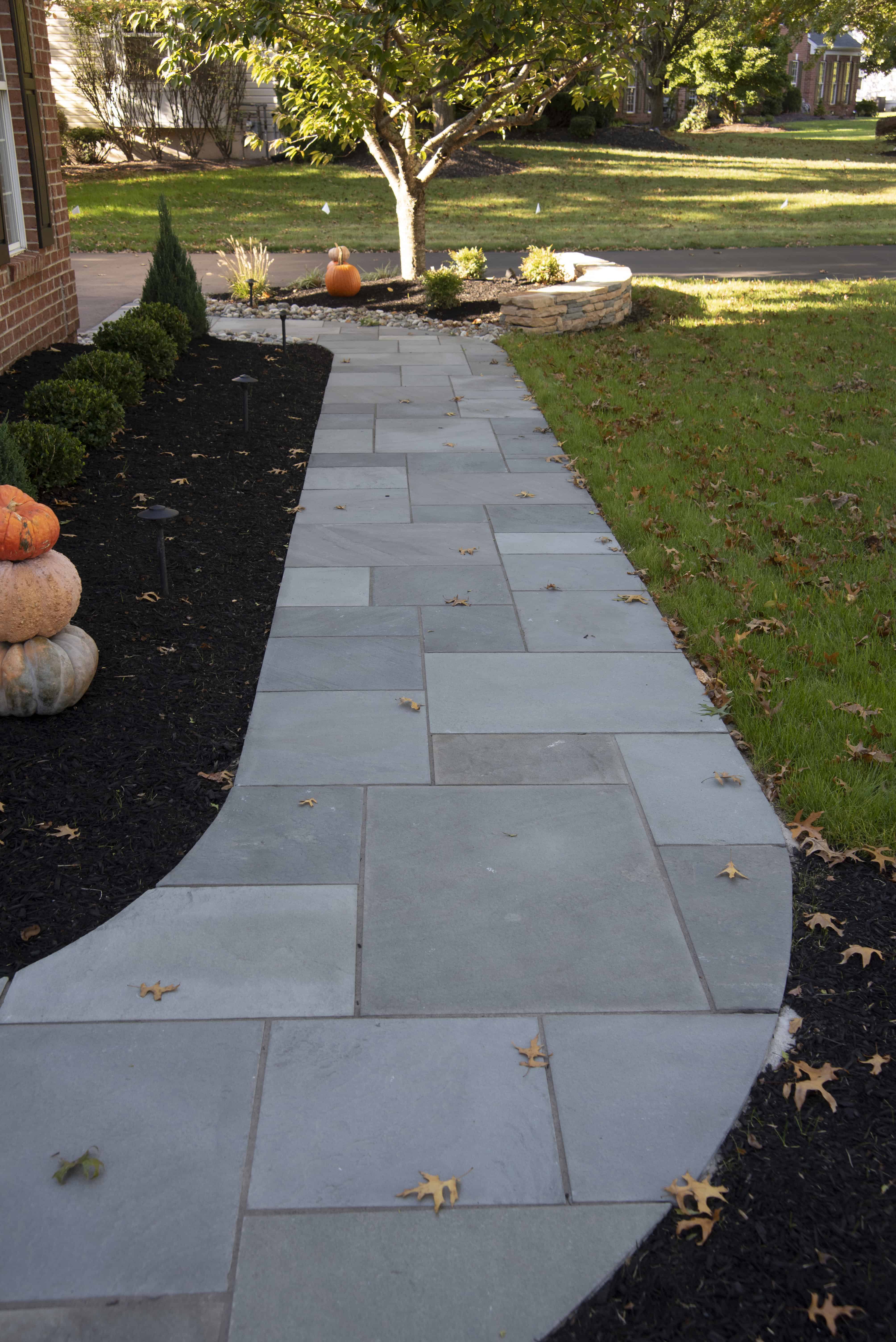 A curved stone walkway bordered by mulch and bushes on one side and grass on the other, with scattered autumn leaves and several stacked pumpkins near a tree in the background.