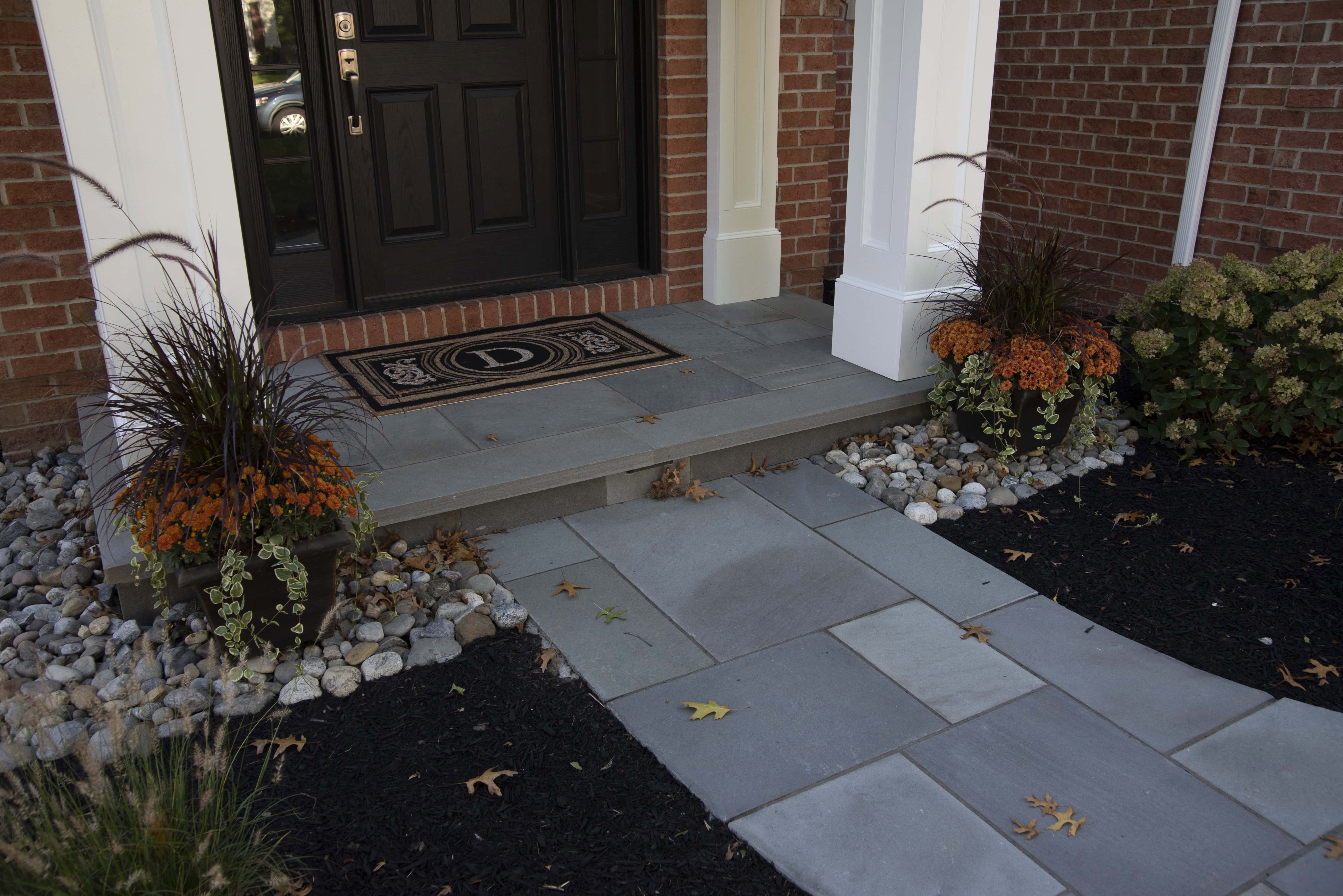 Front porch with a black door, a welcome mat, and two potted plants with orange flowers on either side of the entrance. Stone steps and walkway are bordered by rocks and some fallen leaves.