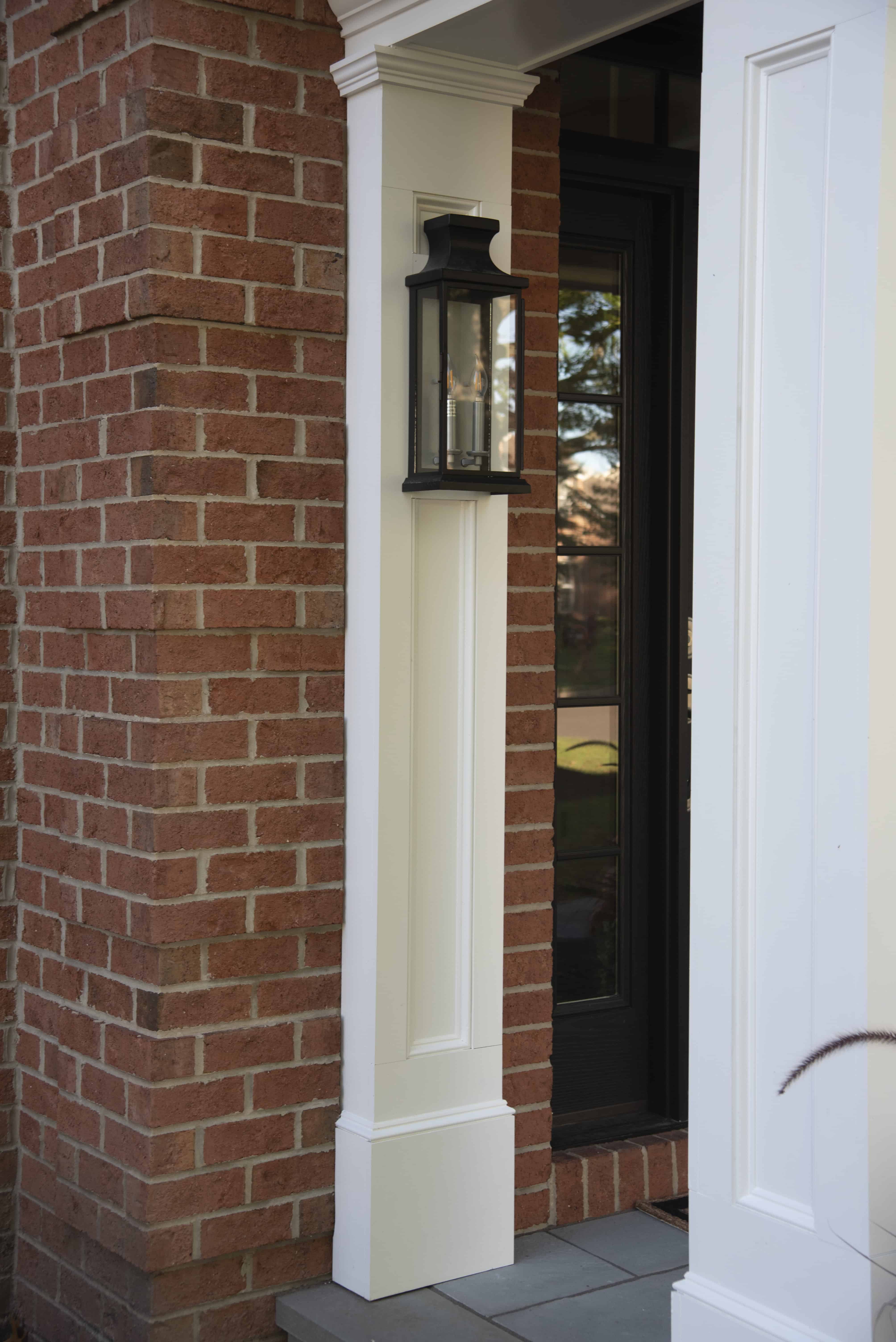 A close-up of a front entrance with red brick walls, a white column, a black outdoor lantern, and a black framed glass door reflecting trees and sky.
