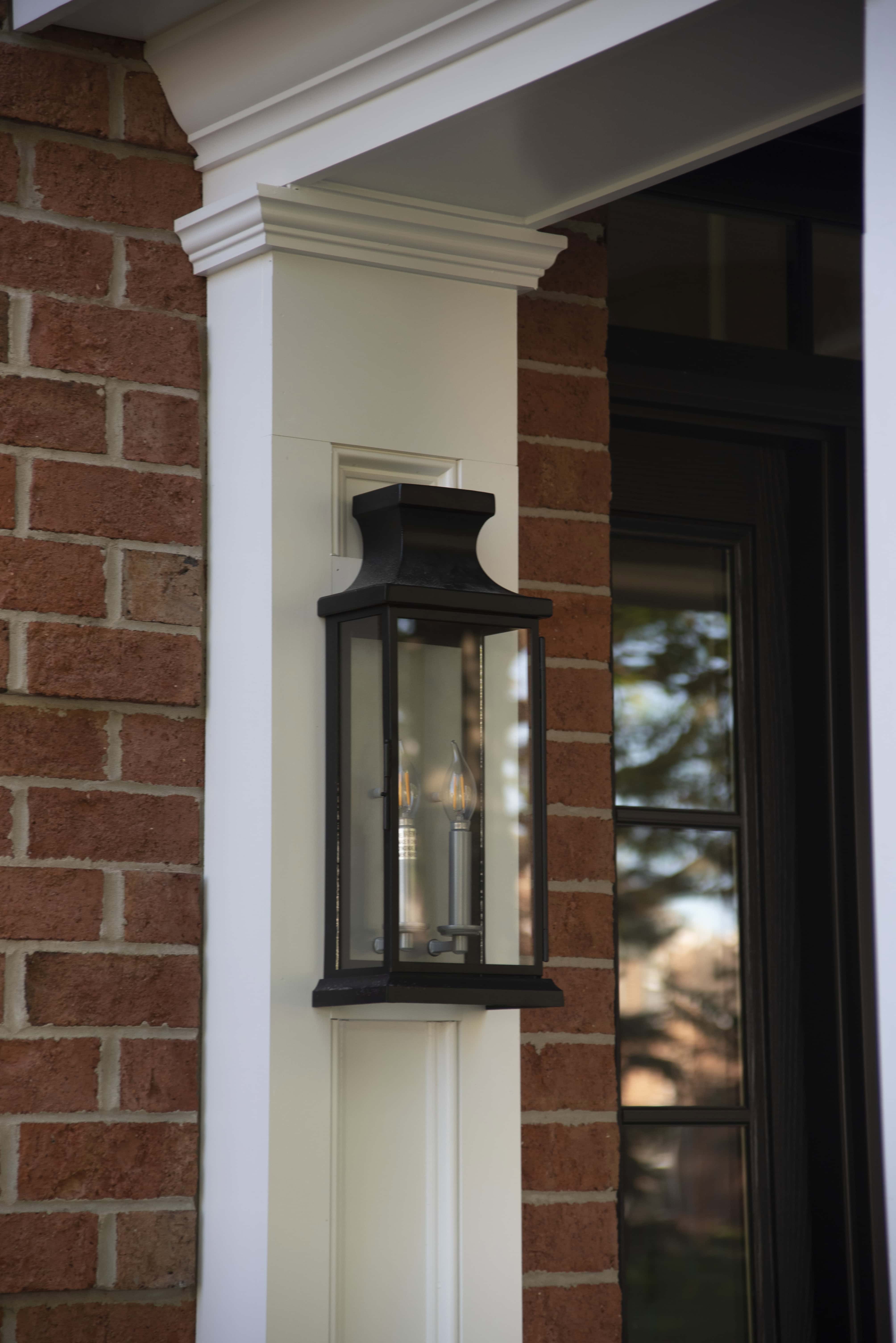 A black metal and glass outdoor wall lantern is mounted on a white trim beside a brick wall and a window with black framing.