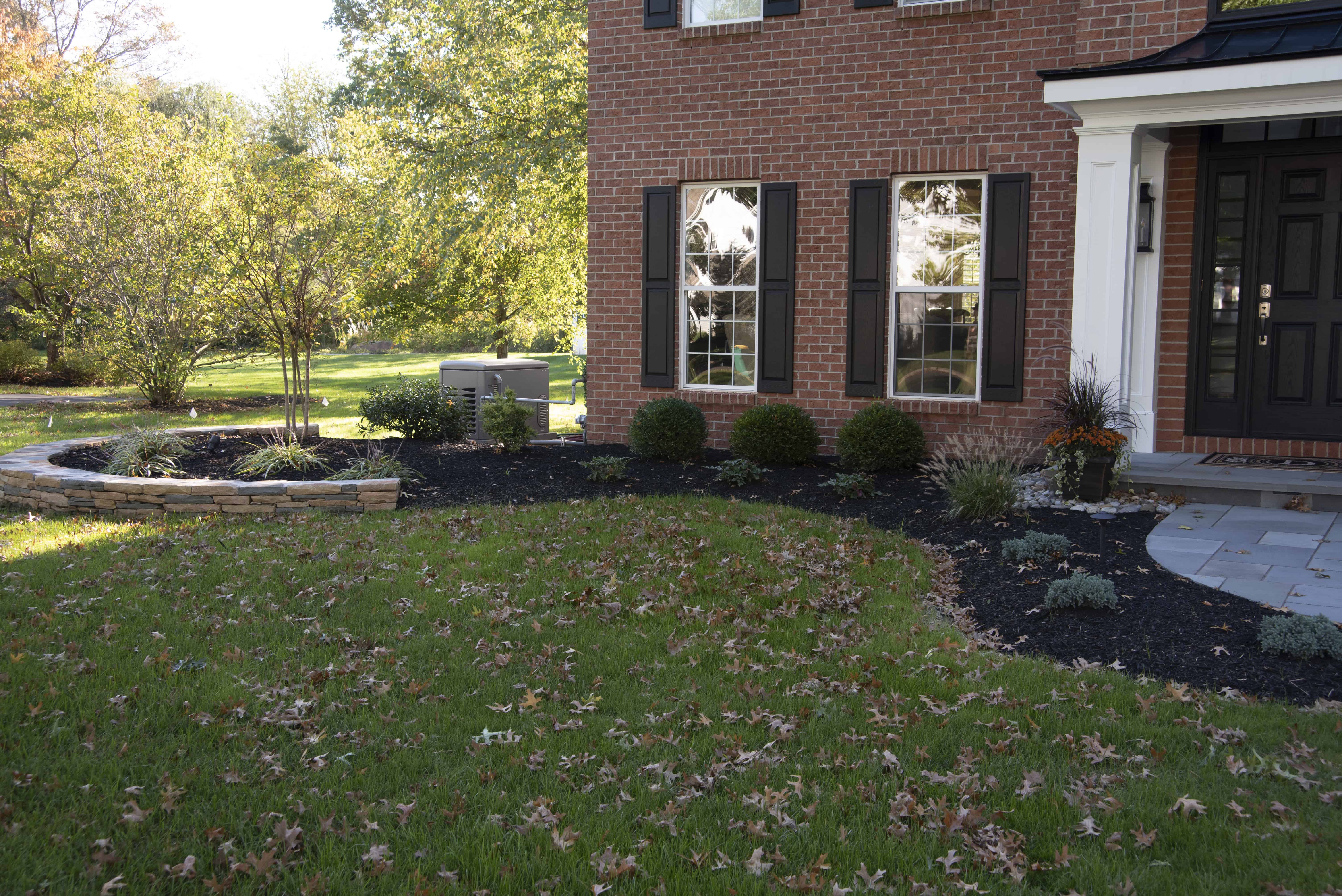 A brick house with black shutters and a black door, surrounded by a landscaped yard. The ground is covered with scattered autumn leaves, green grass, bushes, mulched flower beds, and a low stone wall.