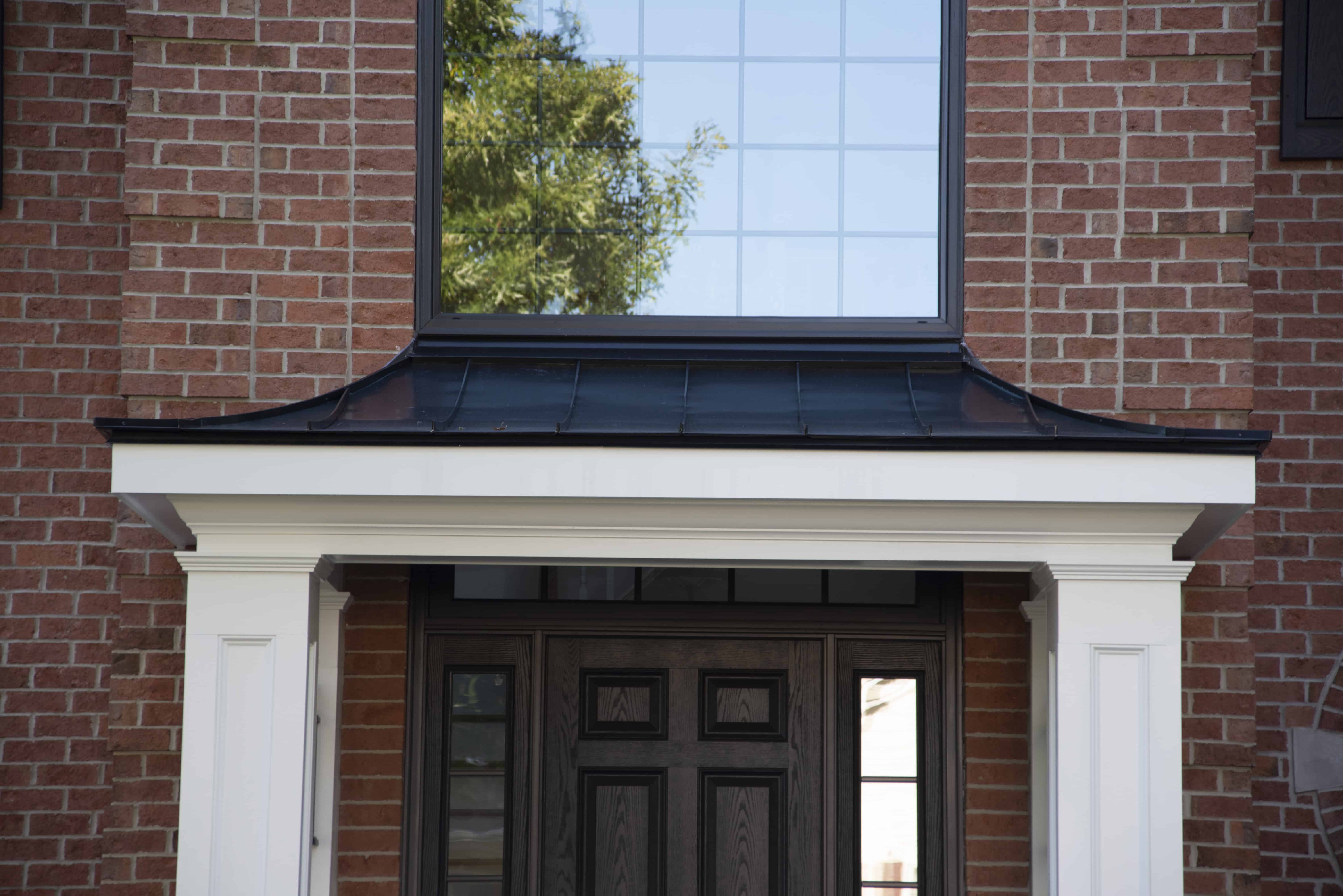 A close-up of a brick house entrance with a dark wooden door, white trim, and a black metal awning. A large window with reflections of trees is above the doorway.