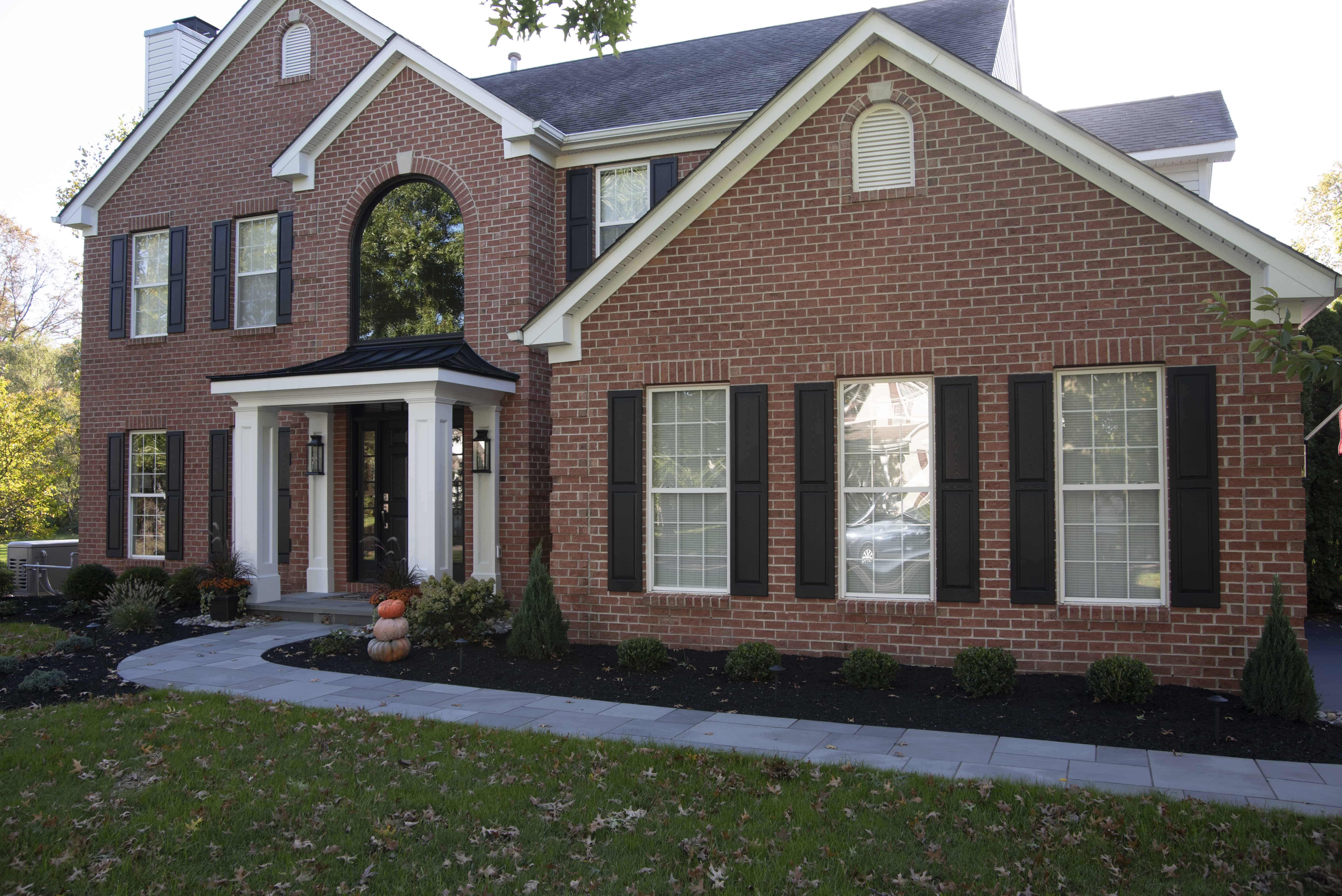 A two-story red brick house with black shutters, white trim, and a front porch decorated with pumpkins. The yard has green grass, shrubs, and a stone walkway leading to the entrance.