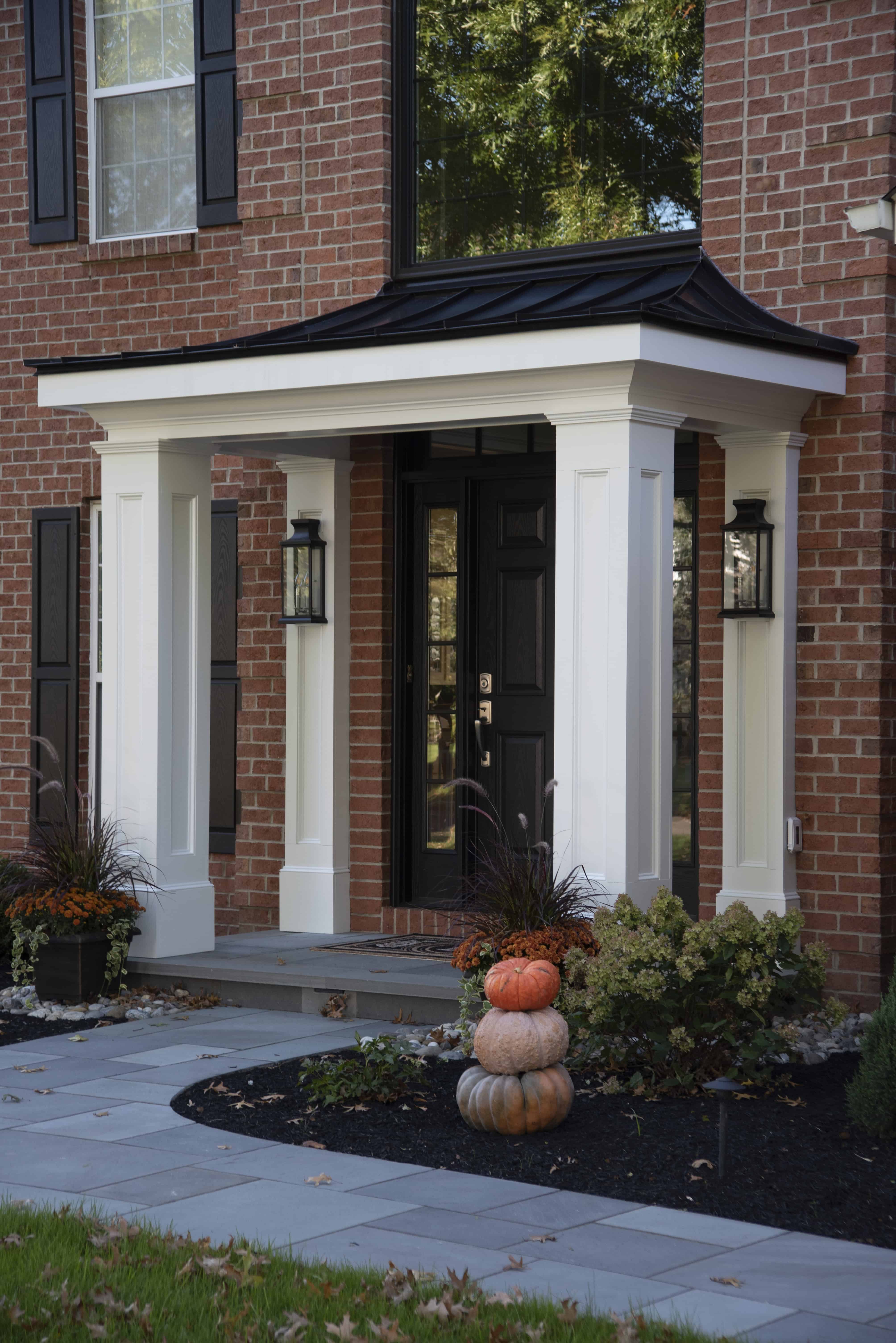 A brick house entryway with white columns, black front door, side lanterns, and a stack of pumpkins on the porch, surrounded by decorative plants and a stone pathway.