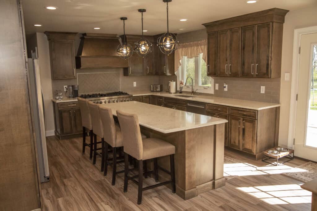 Spacious modern kitchen with wooden cabinets, a large island with four upholstered chairs, pendant lights, stainless steel appliances, and light wood flooring. Sunlight streams in through a glass door.
