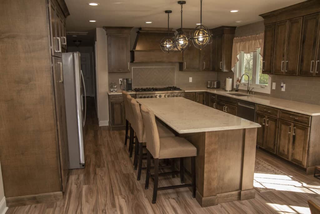 A modern kitchen with wooden cabinets, a large island with three upholstered chairs, stainless steel appliances, pendant lights, and a window above the sink letting in natural light.