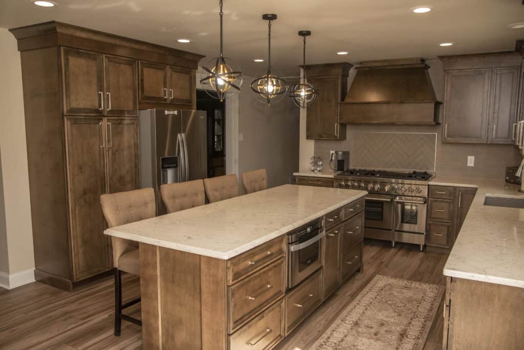 Modern kitchen with wooden cabinets, a large marble island with beige chairs, stainless steel appliances, pendant lights, and a patterned rug on wood flooring. The space is well-lit and organized.