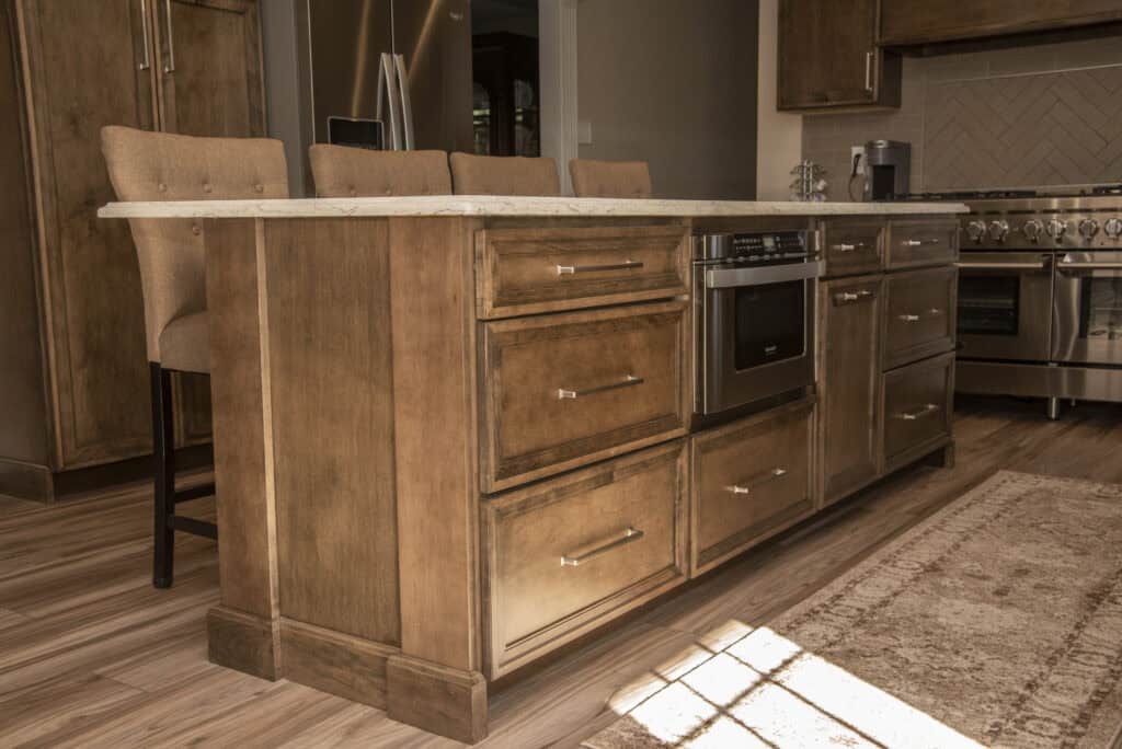A modern kitchen island with brown wooden cabinets, stainless steel handles, a built-in microwave, and a light marble countertop, surrounded by beige upholstered bar stools on a wooden floor.