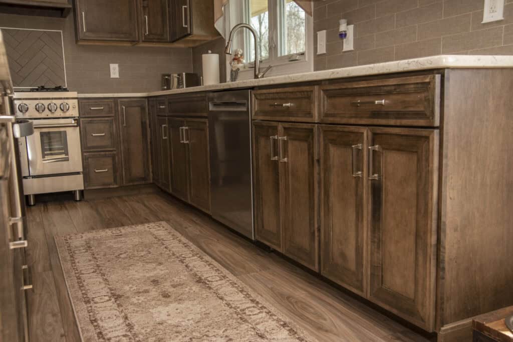 A modern kitchen with dark wooden cabinets, stainless steel appliances, a dishwasher, a light-colored countertop, and a patterned rug on wooden flooring. A window above the sink lets in natural light.