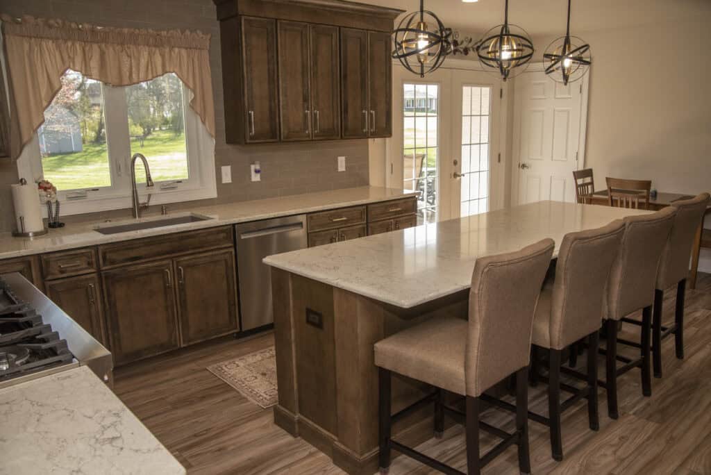 A modern kitchen with brown cabinets, a large island with a marble countertop, four upholstered barstools, stainless steel appliances, and a window overlooking a green yard. Spherical pendant lights hang above the island.