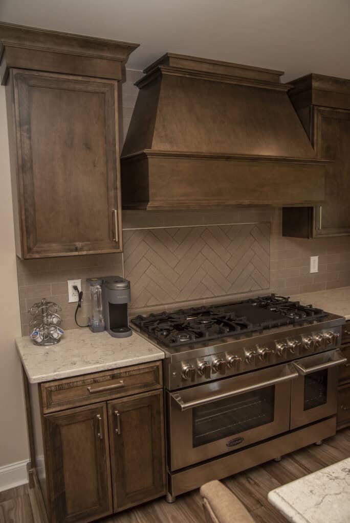 A modern kitchen with wooden cabinets, a stainless steel gas stove and oven, a large wooden range hood, a Keurig coffee maker, and a marble countertop. The backsplash features a herringbone tile pattern.