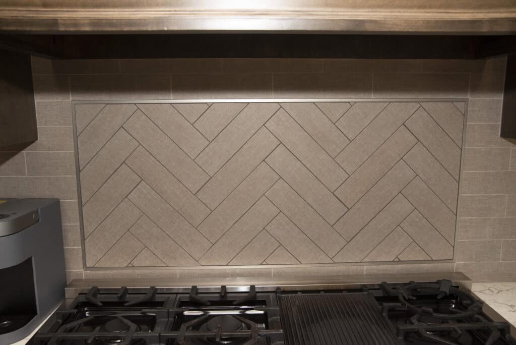 A kitchen backsplash featuring tan tiles arranged in a herringbone pattern, framed by rectangular tiles, with a gas stovetop and metal vent hood in the foreground.