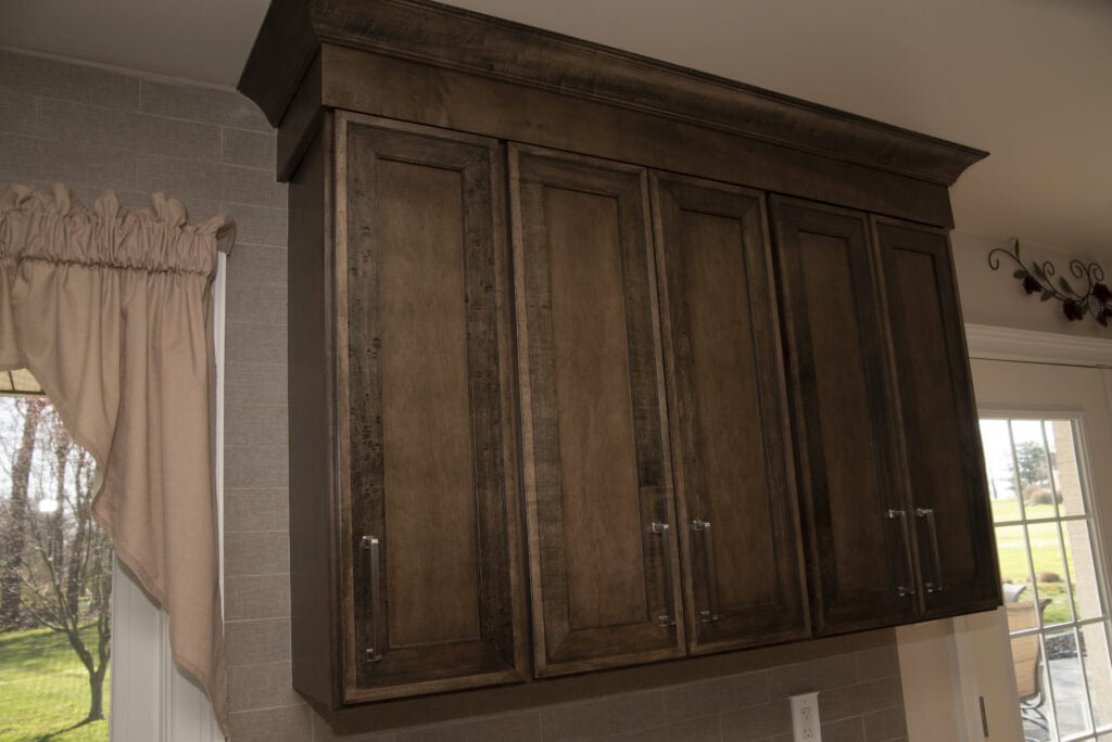 Dark wood kitchen cabinets with four paneled doors and silver handles are mounted on a light-colored tiled wall. A window with a beige curtain and a glass door are visible nearby.