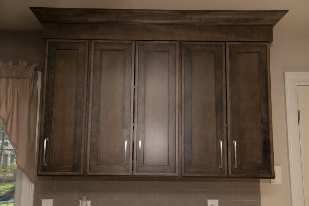 Four tall, dark wood kitchen cabinets with silver handles are mounted on a wall above a light brown tiled backsplash. There is a beige wall, a window with a tan curtain, and a white door in the background.