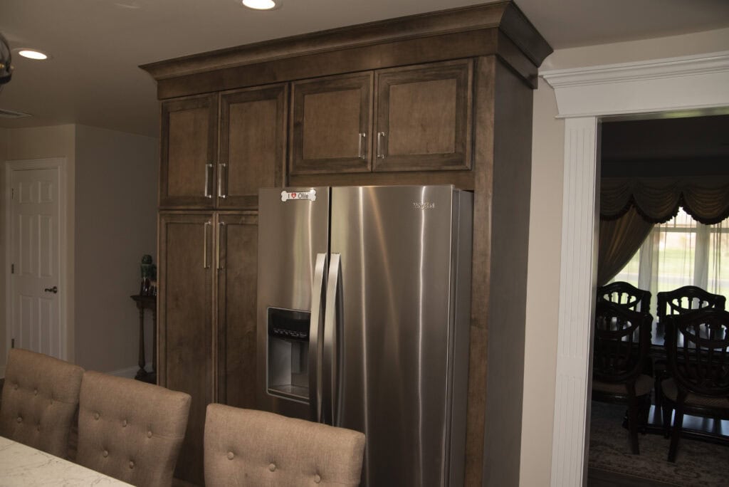 A modern kitchen with brown wooden cabinets, a stainless steel double-door refrigerator with a water dispenser, beige cushioned chairs, and a view into a dining room with a window and curtains.