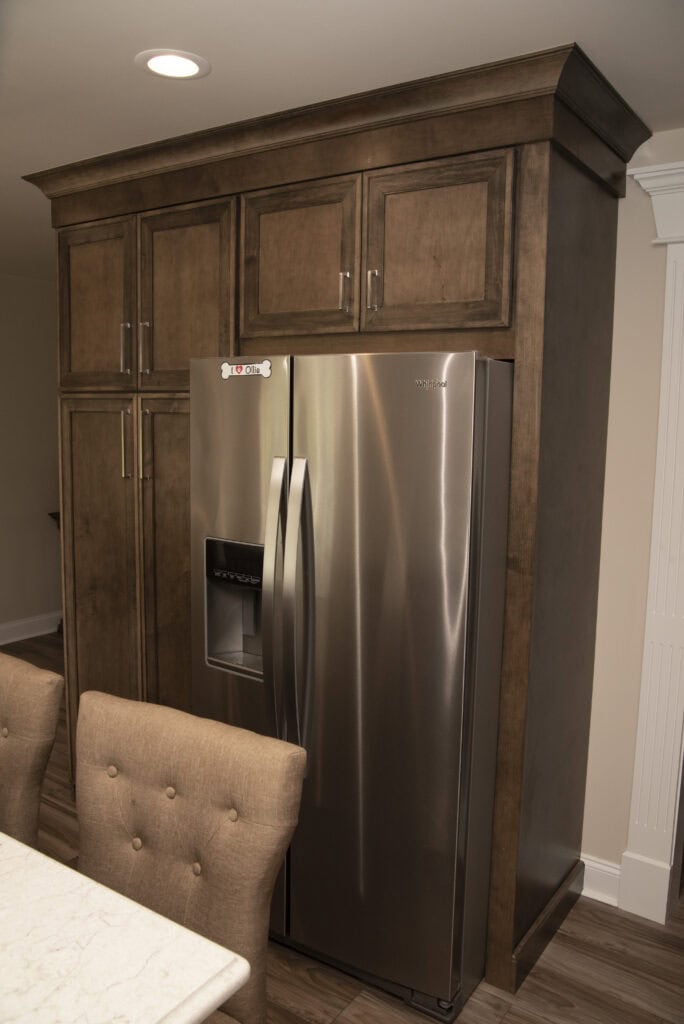 Stainless steel refrigerator with a built-in water and ice dispenser, surrounded by dark wood cabinetry in a modern kitchen. Two upholstered dining chairs are partially visible in the foreground.