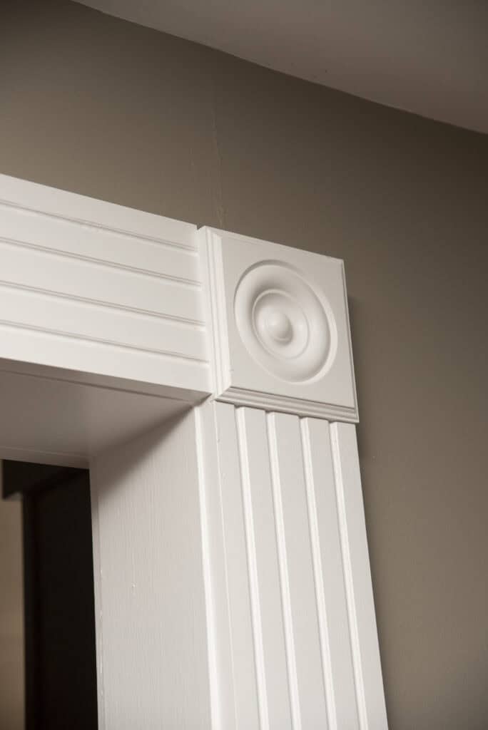 Close-up of a white decorative door trim with vertical grooves and a square rosette detail at the corner, set against a taupe-colored wall.