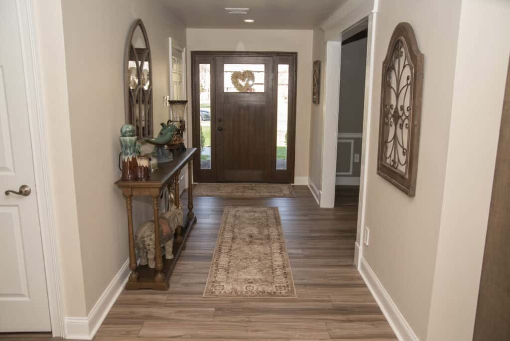 A hallway with wood floors, a patterned rug, a decorative wooden console table with ornaments, a wall mirror, and a wooden front door with glass panels and a heart-shaped wreath.