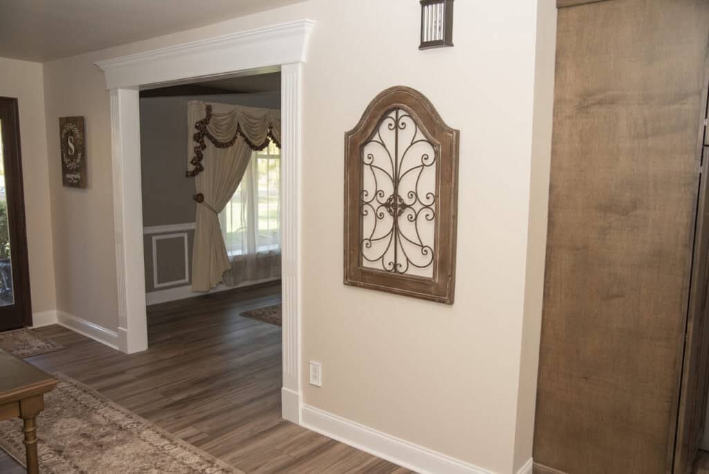 A hallway with light-colored walls and wood flooring, featuring a decorative metal and wood wall art piece. An open doorway leads to a sunlit room with draped curtains and detailed trim.