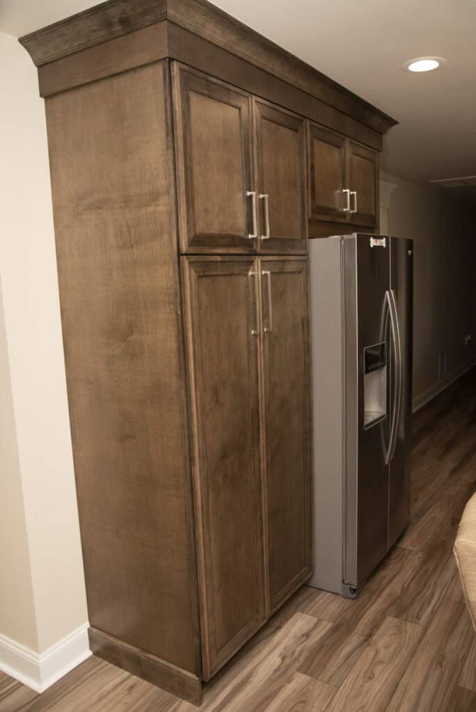 A stainless steel refrigerator is built into wooden kitchen cabinetry with panel doors, handles, and crown molding. The cabinets are medium brown, and the floor is wood. The scene is well-lit.