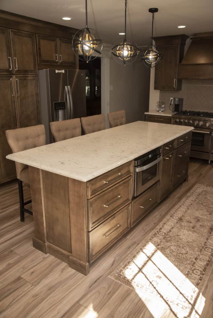 A modern kitchen with wooden cabinets, a marble island with cushioned chairs, stainless steel appliances, pendant lights, and a patterned rug on wood flooring. Sunlight streams onto the island and floor.