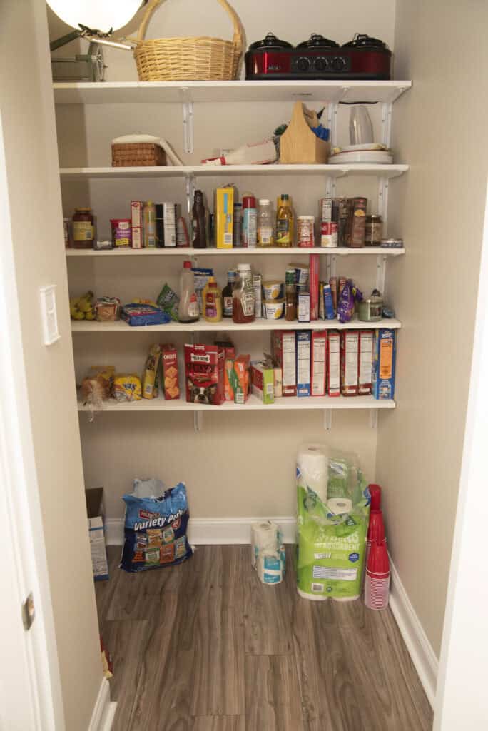 A pantry with shelves holding canned goods, cereal boxes, snacks, sauces, and kitchen items; the floor has paper towels, napkins, red plastic cups, and pet food bags. The floor is wooden, and the walls are beige.