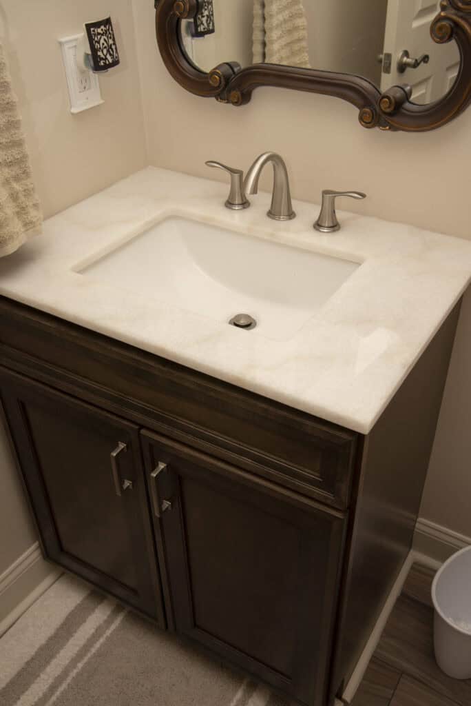 A bathroom vanity with a white countertop, rectangular sink, and brushed nickel faucet. There’s a mirror above, towels on the wall, a striped floor mat, and a trash can is partially visible to the right.