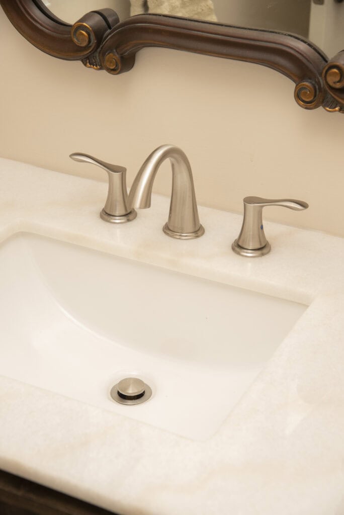 A close-up of a bathroom sink with a brushed nickel faucet and two handles, set in a light-colored countertop with part of a decorative framed mirror visible above.