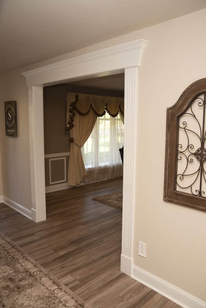 A view through a wide doorway into a room with wood flooring, large curtained windows, beige walls, and elegant decor, including a decorative metal wall hanging and ornate drapes.