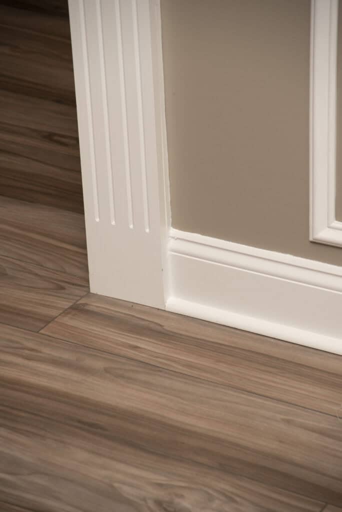 A close-up view of a corner where white-painted trim meets a beige wall and light brown wood-patterned flooring. The trim features vertical grooves, and the floor has a natural wood grain appearance.