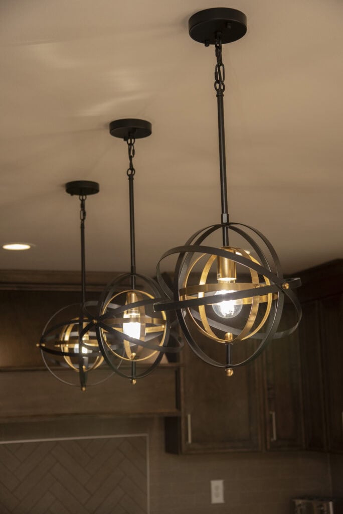 Three modern pendant lights with black and gold spherical metal frames hang from a white ceiling, illuminating a kitchen with dark wood cabinets in the background.